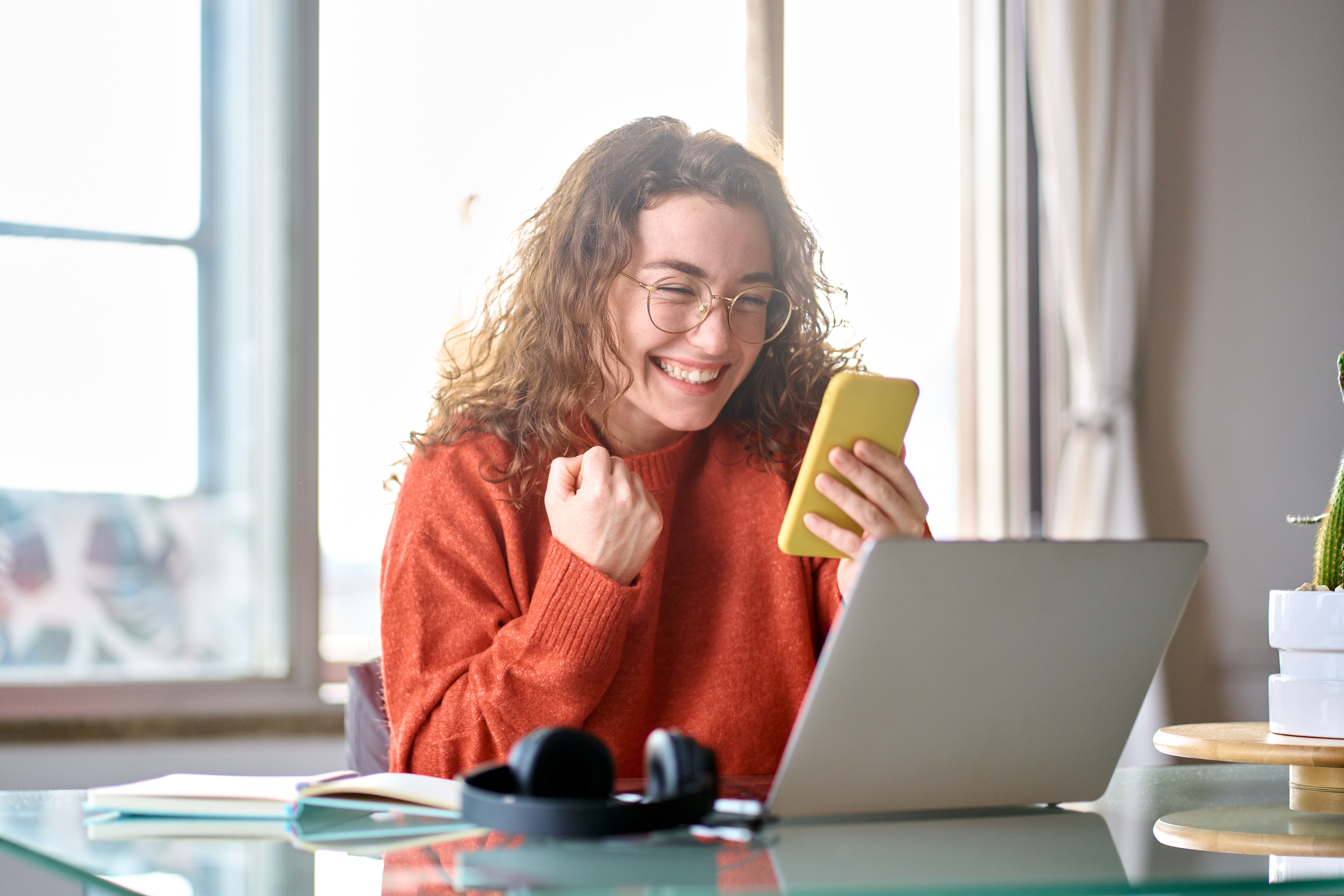 Woman wearing glasses and a red sweater sitting in front of a laptop and holding her phone, pumping her fist victoriously.