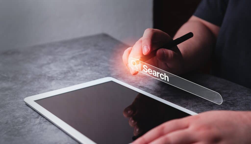 Closeup on a pair of human hands. There is a tablet on the table and one of the hands is holding a stylus and pressing a search box that is hovering in the air above the tablet.